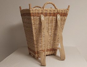 Ein rechteckiger Holztragekorb mit eingeflochtenen Holzstäben an den Ecken und zwei Tragegriffen. Er besteht aus heller, geflochtener Weide in die zwei dunkelbraune Streifen eingeflochten sind. Diese befinden sich in geringem Abstand vom unteren und oberen Rand des Korbes. Er hat zwei breite, beige Tragegurte für schwere Lasten.