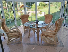 Diese Sitzgruppe steht in einem Wintergarten aus weissen Glastüren und beigen Fliesen. In der Mitte stehen vier geschwungene Rattansessel mit Kopfstützen um einen Tisch mit runder Glasplatte, der ebenfalls aus Rattan hergestellt ist. Auf dem Tisch stehen Kaffeegedecke.