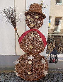 Auf dem gepflasterten Gehweg vor dem Geschäft steht ein geflochtener Schneemann mit einem Hexenbesen und rotem Schal als Aussendekoration. Er hat einen Weidenhut auf und ist mit weissen Augen, Nase und Mund sowie Knöpfen dekoriert. In ihm steckt an einem Holzstab eine schwarze Laterne mit roter Kerze aus Metall.