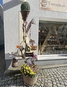 Ebenfalls auf dem gepflasterten Gehweg steht ein verzweigter Birkenstamm in einem geflochenen, runden Blumenkorb. In ihm sind gelbe und lila Stiefmütterchen gepflanzt. An dem Birkenstamm hängen verschiedene geflochtene Artikel. Oben endet er in einem dunklen Weidentrichter, der mit einem Buchsbaum bepflanzt ist.