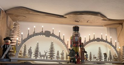 Auf einem Wandregal stehen zwei beleuchtete und geschnitzte Lichterbögen. In ihnen befinden sich geschnitzte Waldtiere und Bäume. Zwischen den Lichterbögen steht ein großer roter Nusskacker gemeinsam mit drei kleineren Räuchermännchen.