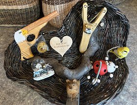 In einer dunklen Schale aus Weidenzweigen sind Holzspielzeug, Schneekugeln und Souvenirs zu sehen. Das Holzspielzeug besteht aus einer kleinen Holzpistole und einer Steinschleuder. Bei den Souvenirs sind ein roter Marienkäfer, ein gelber Vogel und ein kleines hölzernes Herz zu sehen.