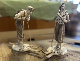 Dieses Bild wird durch zwei hochwertig geschnitzte, nostalgische Skifahrer ausgefüllt. Ein Mann und eine Frau tragen historische Skikleidung, hölzerne Ski und Bambusstöcke mit Weidenringen und Lederriemen.