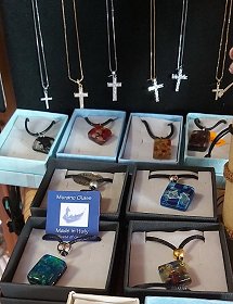 Murano Glasschmuck aus Italien ist auf diesem Bild dargestellt. Silberne Ketten mit funkelnden Christus-Kreuzen oder farbige Schächtelchen mit bunten Glasanhängern und dunklen Lederriemchen zeigen verschiedene Facetten dieses Schmuckherstellers.