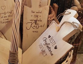 Hölzerne Schneid- und Dekobretter mit verschiedenen Sprüchen liegen in einem hellen Weidenkorb mit Henkel. Ein dunkelbrauner Spruch lautet zum Beispiel - Beste Großeltern der Welt, umrahmt von zwei Herzen.